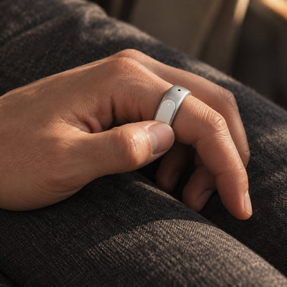 A close-up of a person's hand wearing a slim, light gray smart ring, resting on their dark gray trousers.